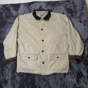 Könnte beinhalten: Eine beigefarbene Langarmjacke mit einem dunkelbraunen Cordkragen und passenden Cordbündchen. Die Jacke verfügt über eine Knopfleiste, zwei untere Vordertaschen mit Knopfklappen sowie zwei schräge Brusttaschen.