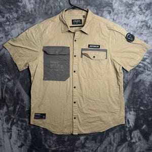 May include: A tan short-sleeved button-down shirt featuring a dark gray flap pocket on the left chest with printed text, a smaller right chest pocket with an AVIREX label, a circular patch on the left sleeve, and a small rectangular label at the hem.