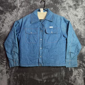 May include: A blue denim jacket featuring a cream-colored sherpa lining, a pointed collar, and a front button closure. The jacket includes two chest pockets with button flaps and long sleeves with buttoned cuffs, hanging on a black plastic hanger.
