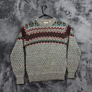 May include: A long-sleeve crew neck jumper in a heathered grey knit. The upper chest features a horizontal band with a geometric pattern in dark red, forest green, and cream. The remainder of the jumper has a small, repeating green dot pattern.