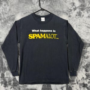 May include: A black long-sleeve t-shirt featuring the white text "What happens in" above the yellow text "SPAMALOT..." printed on the chest. The shirt has a crew neckline and is displayed on a black hanger against a gray tie-dye background.