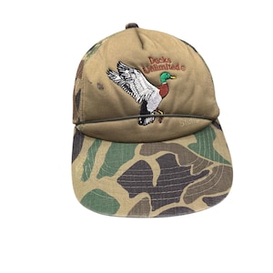 Puede incluir: Una gorra tipo camionero de color tostado con visera de camuflaje y paneles traseros de malla. El panel frontal muestra un pato real bordado en vuelo con el texto Ducks Unlimited en hilo marrón. Un cordón oscuro cruza la base de la corona.