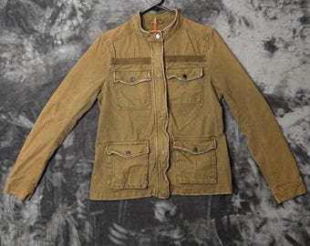 May include: An olive green utility jacket featuring a stand-up collar, a full-length front button closure, and four front patch pockets with buttoned flaps. The jacket has long sleeves and a horizontal fabric panel detail across the chest area.