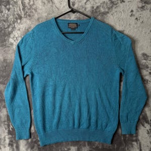 May include: A solid teal blue long-sleeve sweater with a V-neckline and ribbed cuffs and hem. The garment features a Pendleton brand label stitched inside the collar area. The knit texture is consistent throughout the front of the sweater.