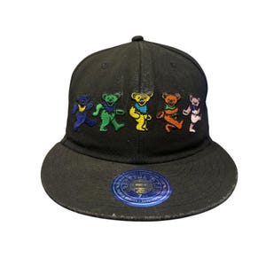 May include: A black baseball cap featuring an embroidered row of five colorful dancing bears in blue, green, yellow, orange, and pink. A circular blue sticker with the text Grateful Dead and the size 7 1/2 is affixed to the flat brim.