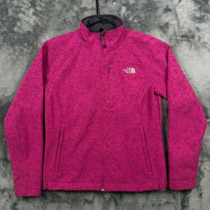 May include: A bright pink long-sleeve jacket featuring a black geometric spiderweb-like pattern throughout. The jacket has a full-length front zipper, a high collar, and a small white The North Face logo embroidered on the upper left chest area.