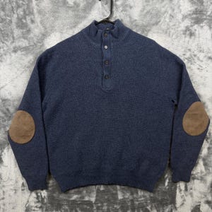 Op de afbeelding: Een marineblauwe trui met lange mouwen, voorzien van een opstaande kraag met een knoopsluiting van vier knopen. Het kledingstuk is gemaakt van een gebreide stof met textuur en heeft contrasterende bruine elleboogstukken van suèdine.