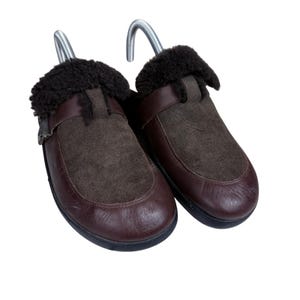 May include: A pair of dark brown leather and suede slip-on shoes featuring a dark brown faux fur lining around the collar and a metal shoe tree insert in each shoe. The shoes have a rounded toe design and a smooth leather finish on the lower section.