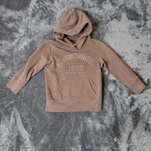May include: A dusty brown long-sleeve hooded sweatshirt featuring a front kangaroo pocket and white embroidered text that reads abercrombie 1892. The garment has ribbed cuffs and a ribbed hem, with a hood that includes a visible interior lining.