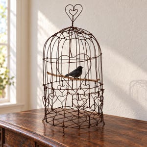 Puede incluir: Una jaula decorativa para pájaros hecha de alambre de color marrón oscuro oxidado, con una parte superior en forma de cúpula y un remate en forma de corazón. La estructura de la jaula incorpora múltiples detalles de alambre en forma de corazón y contiene un pequeño pájaro negro posado en una rama de madera fina en su interior.