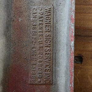 Puede incluir: Primer plano de un letrero met&aacute;lico desgastado con el texto "WAGNER SIGN SERVICES INC." e informaci&oacute;n de patentes. El letrero tiene un borde rojo y una apariencia texturizada y envejecida, lo que sugiere que es vintage.