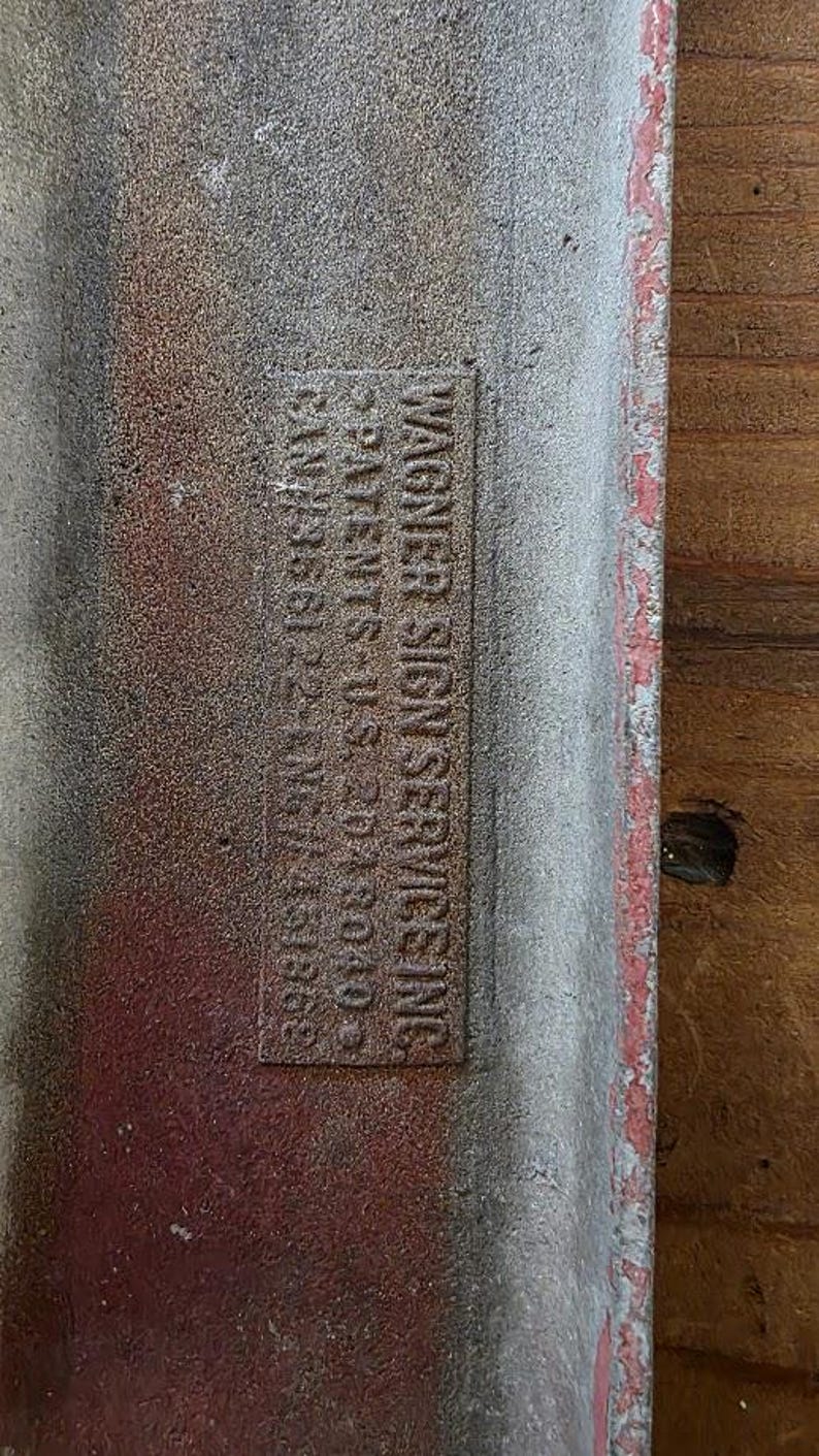 Puede incluir: Primer plano de un letrero met&aacute;lico desgastado con el texto "WAGNER SIGN SERVICES INC." e informaci&oacute;n de patentes. El letrero tiene un borde rojo y una apariencia texturizada y envejecida, lo que sugiere que es vintage.