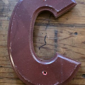 Puede incluir: Una letra "C" de madera de color granate, con un acabado envejecido. La letra tiene una forma redondeada con una parte superior e inferior planas. Cuenta con dos peque&ntilde;os agujeros para colgar y est&aacute; ensartada con hilo.