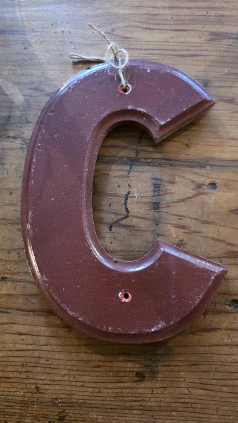 Puede incluir: Una letra "C" de madera de color granate, con un acabado envejecido. La letra tiene una forma redondeada con una parte superior e inferior planas. Cuenta con dos peque&ntilde;os agujeros para colgar y est&aacute; ensartada con hilo.