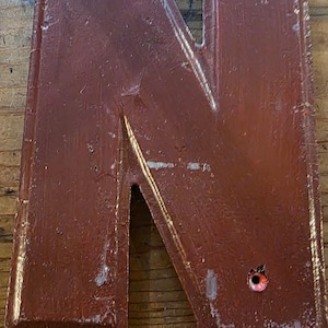 Puede incluir: Una gran letra "N" de color burdeos desgastado, con una est&eacute;tica r&uacute;stica y vintage. La letra de madera tiene un efecto de pintura texturizada y descascarillada y est&aacute; ensartada con hilo para colgar. La letra se muestra sobre una superficie de madera.