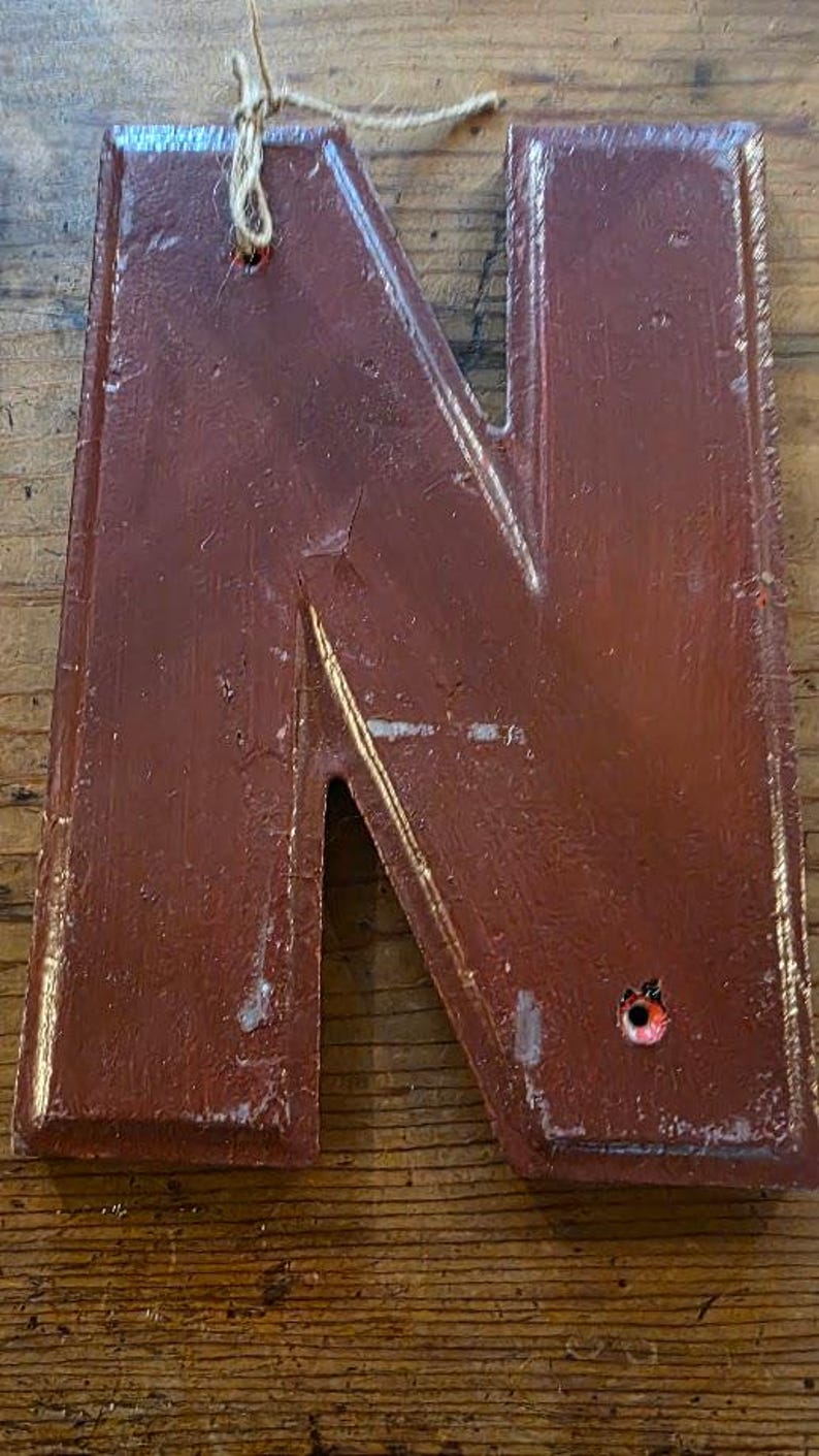 Puede incluir: Una gran letra "N" de color burdeos desgastado, con una est&eacute;tica r&uacute;stica y vintage. La letra de madera tiene un efecto de pintura texturizada y descascarillada y est&aacute; ensartada con hilo para colgar. La letra se muestra sobre una superficie de madera.