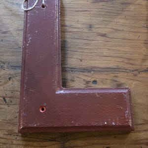 Puede incluir: Una letra "L" de madera marr&oacute;n con un acabado envejecido. La letra tiene un peque&ntilde;o agujero cerca de la parte superior y un lazo de cuerda para colgar. El fondo es una superficie de madera.