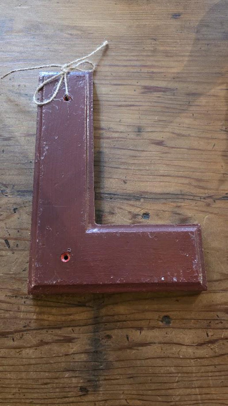 Puede incluir: Una letra "L" de madera marr&oacute;n con un acabado envejecido. La letra tiene un peque&ntilde;o agujero cerca de la parte superior y un lazo de cuerda para colgar. El fondo es una superficie de madera.
