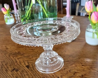 Könnte beinhalten: Ein klarer Kuchenständer aus Glas mit dekorativem, gezahntem Rand. Der Ständer hat einen runden Sockel und einen zentralen Stiel und wird auf einem Holztisch präsentiert. Der Ständer hat einen Durchmesser von etwa 25 cm.