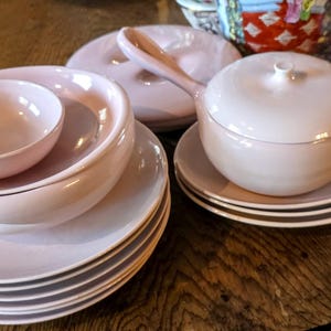 Puede incluir: Una colección de vajilla de cerámica rosa pálido, que incluye platos, cuencos y una fuente con tapa. El conjunto está dispuesto sobre una mesa de madera, mostrando una estética de comedor coherente y elegante. Las piezas tienen un acabado brillante.
