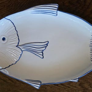 以下が含まれることがあります： 魚の形をした白い陶磁器製の盛り付け皿で、頭部とヒレにエンボス加工のディテールが施されています。ヒレ、尾、魚の目の縁は深い青色の釉薬で縁取られており、白い表面に対してはっきりとしたコントラストを生み出しています。