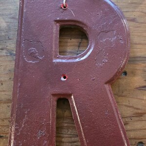 Puede incluir: Una letra "R" de metal granate con un acabado envejecido, con un recorte en el centro y un peque&ntilde;o agujero en la parte superior para colgar. Se adjunta un cordel natural para exhibir. La letra es probablemente para la decoraci&oacute;n del hogar.
