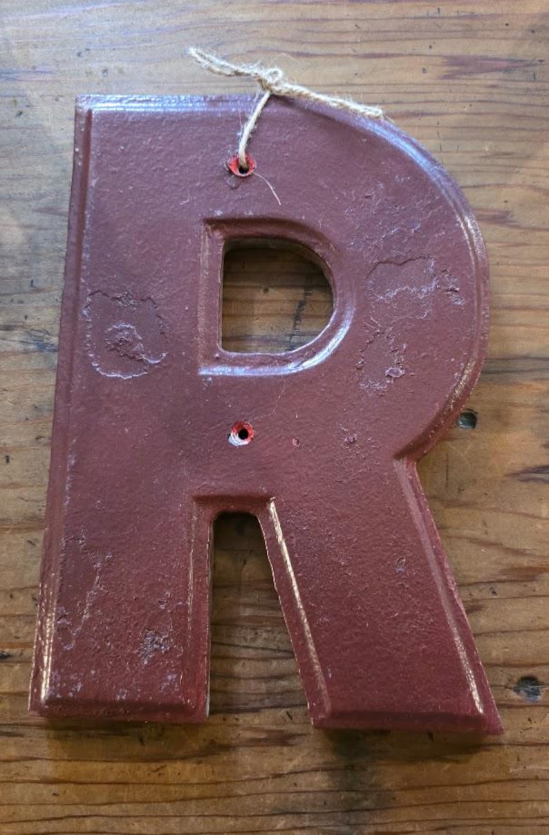 Puede incluir: Una letra "R" de metal granate con un acabado envejecido, con un recorte en el centro y un peque&ntilde;o agujero en la parte superior para colgar. Se adjunta un cordel natural para exhibir. La letra es probablemente para la decoraci&oacute;n del hogar.