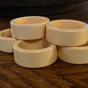 May include: A collection of five ivory-colored napkin rings. Each ring is cylindrical with a rounded exterior and a hollow interior. The rings are stacked and arranged on a dark wooden surface, creating a simple, elegant display.