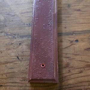 Puede incluir: Una placa rectangular de madera de color granate con una superficie texturizada y un peque&ntilde;o agujero cerca de la parte inferior. Un lazo de cuerda natural est&aacute; atado en la parte superior. La placa mide unos 25 cm de largo.