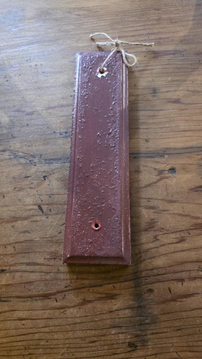Puede incluir: Una placa rectangular de madera de color granate con una superficie texturizada y un peque&ntilde;o agujero cerca de la parte inferior. Un lazo de cuerda natural est&aacute; atado en la parte superior. La placa mide unos 25 cm de largo.