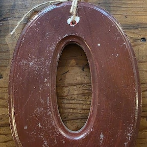 Puede incluir: Un n&uacute;mero "0" decorativo de pared de forma ovalada, en un tono marr&oacute;n rojizo oscuro. El n&uacute;mero tiene un acabado envejecido, con desgaste visible. Se adjunta un lazo de cuerda natural para colgar. Fondo de madera.