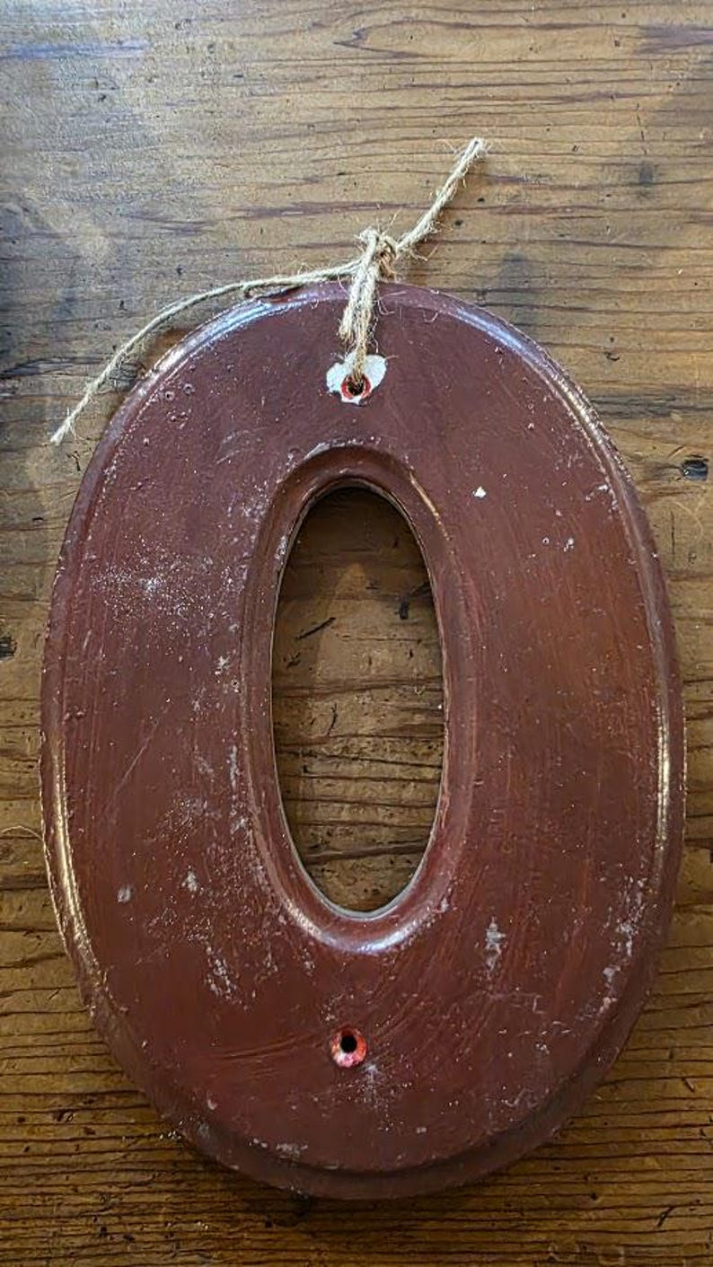 Puede incluir: Un n&uacute;mero "0" decorativo de pared de forma ovalada, en un tono marr&oacute;n rojizo oscuro. El n&uacute;mero tiene un acabado envejecido, con desgaste visible. Se adjunta un lazo de cuerda natural para colgar. Fondo de madera.