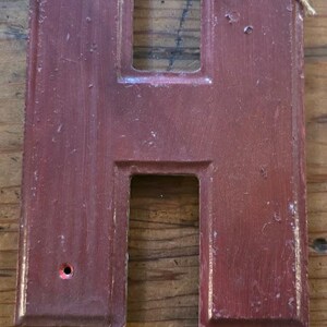 Puede incluir: Letra "H" de madera de color granate, con un acabado envejecido. La letra tiene forma rectangular con un hueco en el centro y un peque&ntilde;o agujero. Un lazo de cuerda est&aacute; sujeto en la parte superior para colgar.