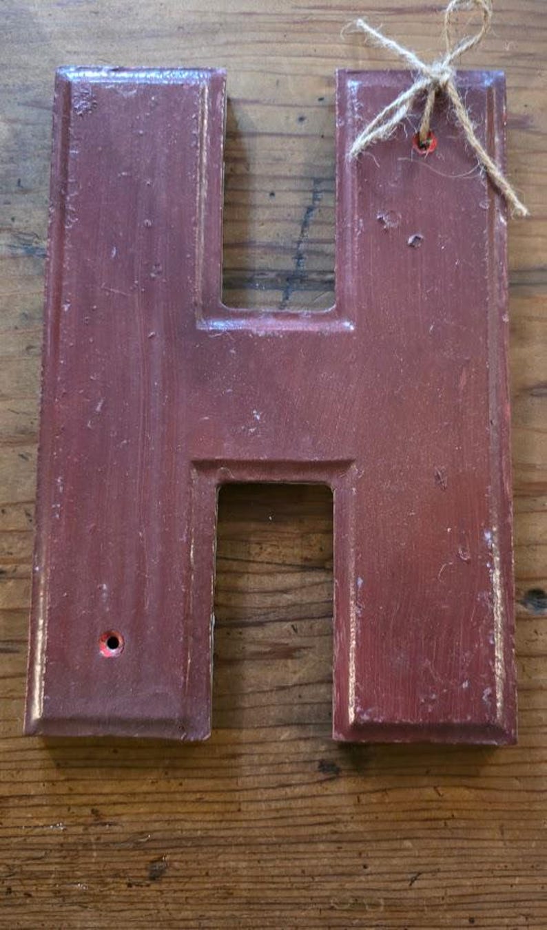 Puede incluir: Letra "H" de madera de color granate, con un acabado envejecido. La letra tiene forma rectangular con un hueco en el centro y un peque&ntilde;o agujero. Un lazo de cuerda est&aacute; sujeto en la parte superior para colgar.