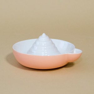 May include: A pink ceramic citrus juicer with a white, cone-shaped reamer. The juicer has a small handle on the side.