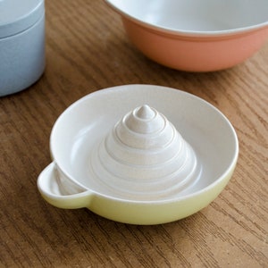 May include: A white ceramic lemon squeezer with a yellow rim. The squeezer has a conical shape with a series of concentric rings.