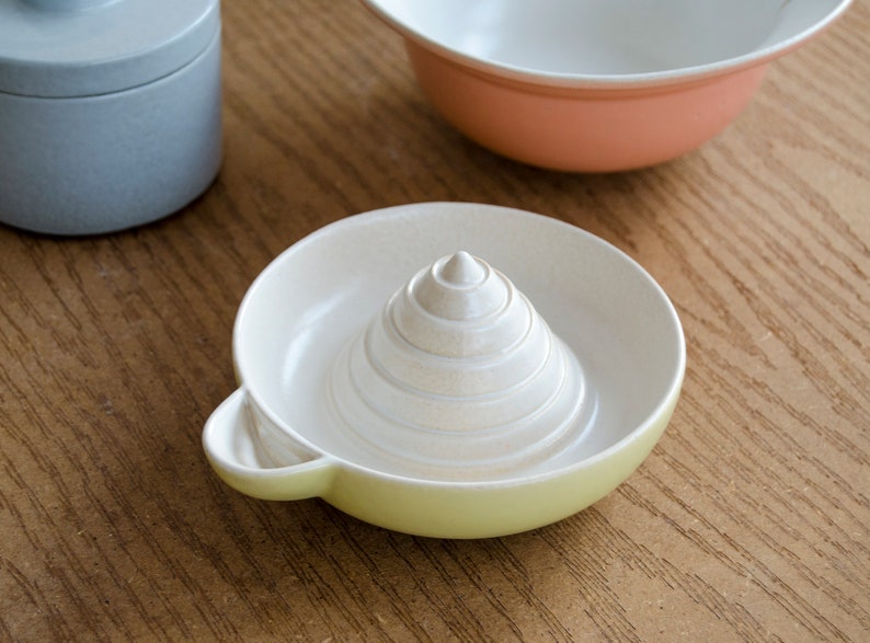 May include: A white ceramic lemon squeezer with a yellow rim. The squeezer has a conical shape with a series of concentric rings.