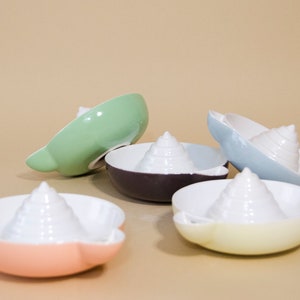 May include: Five ceramic citrus juicers in different colors. The juicers are shaped like seashells with a pointed top and a wide base. The colors are green, purple, light blue, yellow, and pink.