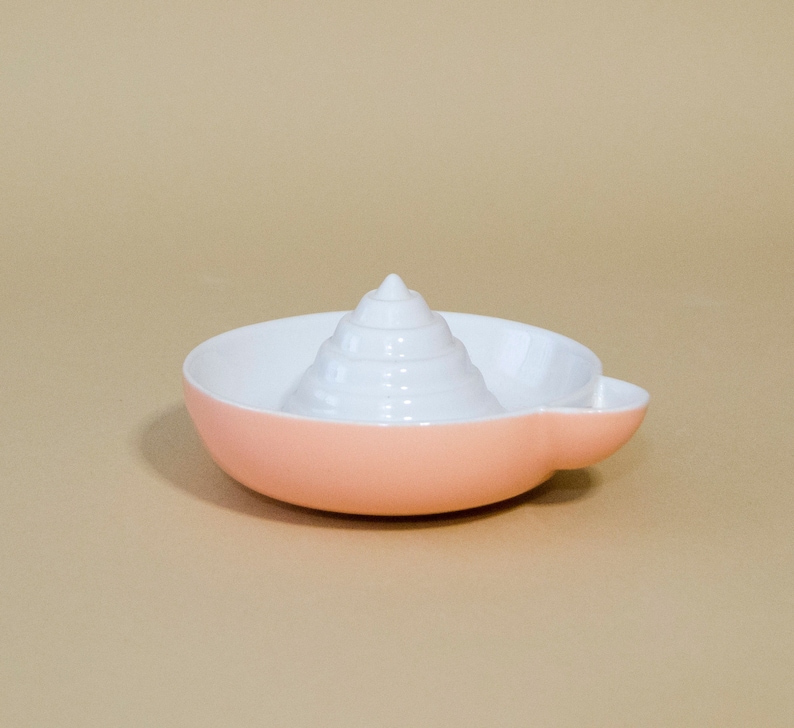 May include: A pink ceramic citrus juicer with a white, cone-shaped reamer. The juicer has a small handle on the side.