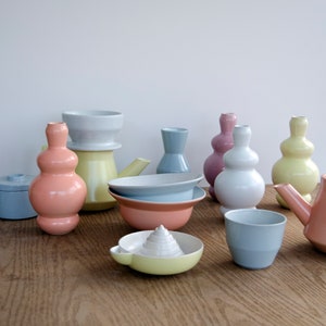 May include: A collection of pastel-colored ceramic vases, bowls, and a teapot. The pieces are arranged on a wooden surface. The colors include light blue, pink, yellow, and white.