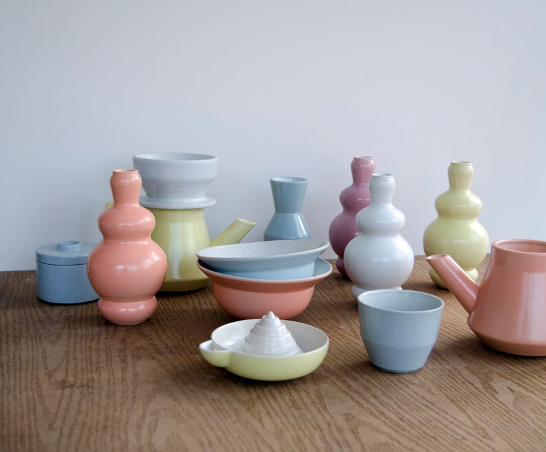 May include: A collection of pastel-colored ceramic vases, bowls, and a teapot. The pieces are arranged on a wooden surface. The colors include light blue, pink, yellow, and white.