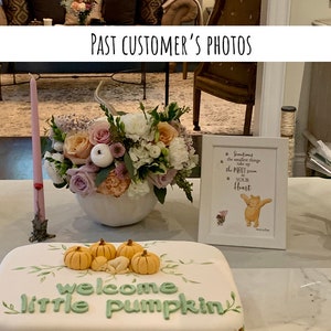 May include: A white cake decorated with a green welcome little pumpkin message and small orange pumpkins. A white vase with pink and white flowers sits on the table next to the cake. A framed Winnie the Pooh quote sits on the table next to the cake.