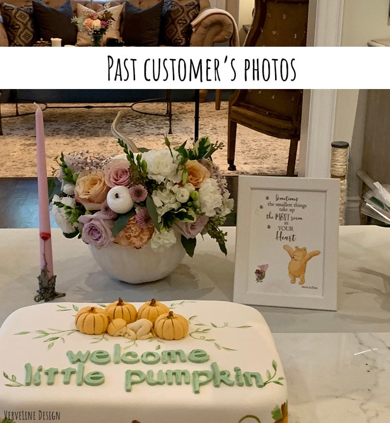 May include: A white cake decorated with a green welcome little pumpkin message and small orange pumpkins. A white vase with pink and white flowers sits on the table next to the cake. A framed Winnie the Pooh quote sits on the table next to the cake.