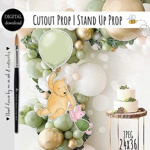 May include: Printable cutout prop featuring Winnie the Pooh and Piglet holding onto a green balloon. The prop is designed to be used as a stand-up prop for a party or event. The image also shows a cake and a balloon garland in the background.