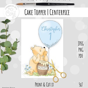 May include: A printable cake topper featuring a cartoon Winnie the Pooh character holding a honey pot. The topper also includes a blue balloon with the name "Christopher" and the number "1" written on it. The topper is designed to be printed and cut out for use as a cake decoration.