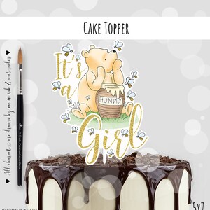 Könnte beinhalten: Ein "Es ist ein Mädchen"-Kuchentopper mit einem Cartoon von Winnie Puuh, der einen Honigtopf hält, um den herum Bienen fliegen. Der Text "Es ist ein Mädchen" ist in goldenen Glitzer geschrieben.