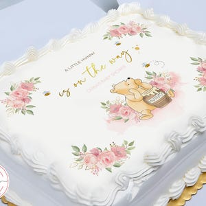 May include: A white cake with a Winnie the Pooh themed design. The cake is decorated with pink flowers, bees, and a honey pot. The text on the cake reads "A little hunny is on the way".
