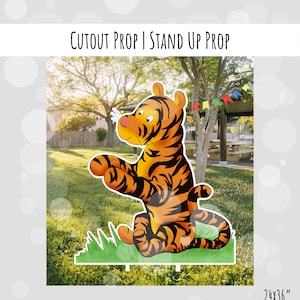 May include: A cutout prop of a cartoon tiger sitting on a patch of green grass. The tiger is orange and black with a white face and blue eyes. The prop is 24 inches by 36 inches.
