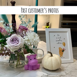 May include: A white framed print with a Winnie the Pooh quote, "It's so much friendlier with two." The print is on a white countertop with a purple hippopotamus toy, a white pumpkin, and a vase of flowers.