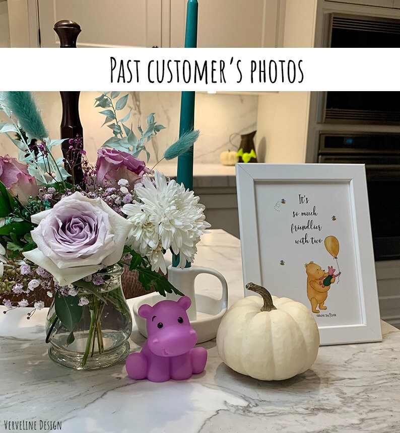 May include: A white framed print with a Winnie the Pooh quote, "It's so much friendlier with two." The print is on a white countertop with a purple hippopotamus toy, a white pumpkin, and a vase of flowers.